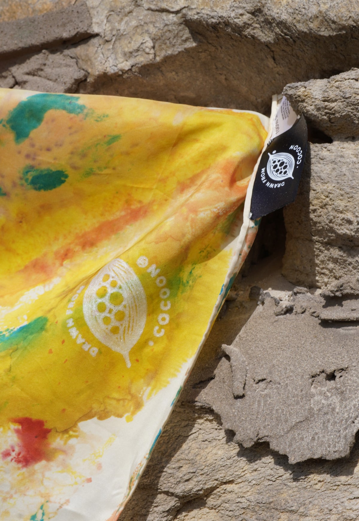 Close-up of a hand-painted silk scarf with the log, in yellow, orange and green, draped over a textured stone wall.