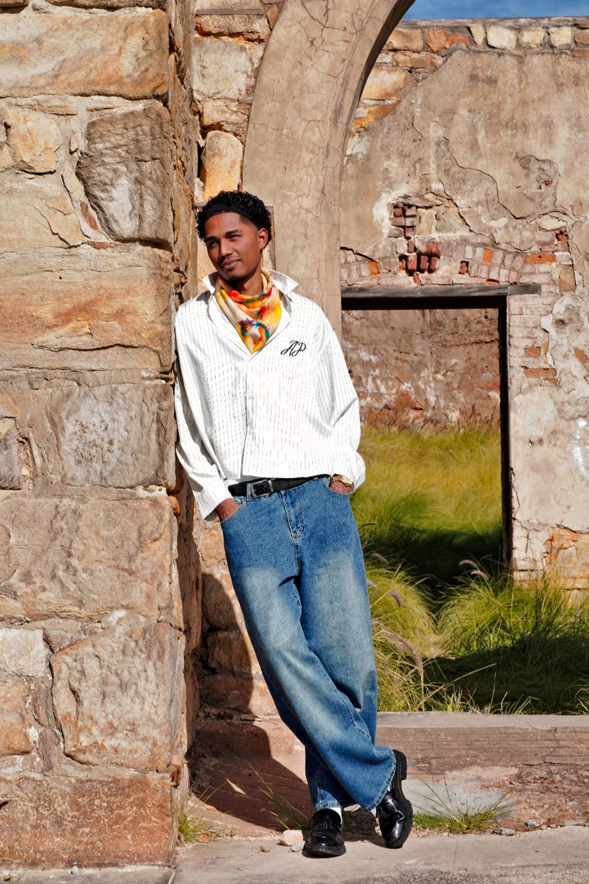 Hand-painted silk scarf worn as a neck scarf or a cravat by a man, photographed against a rustic stone and brick setting.
