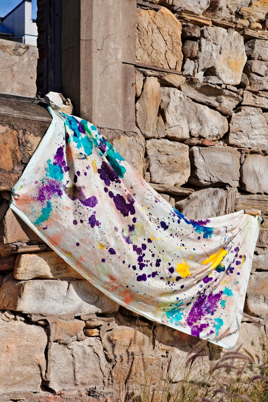 Hand-painted silk wrap draped on a brick wall, showcasing an abstract pattern in purple, blue, green, yellow, coral and red.