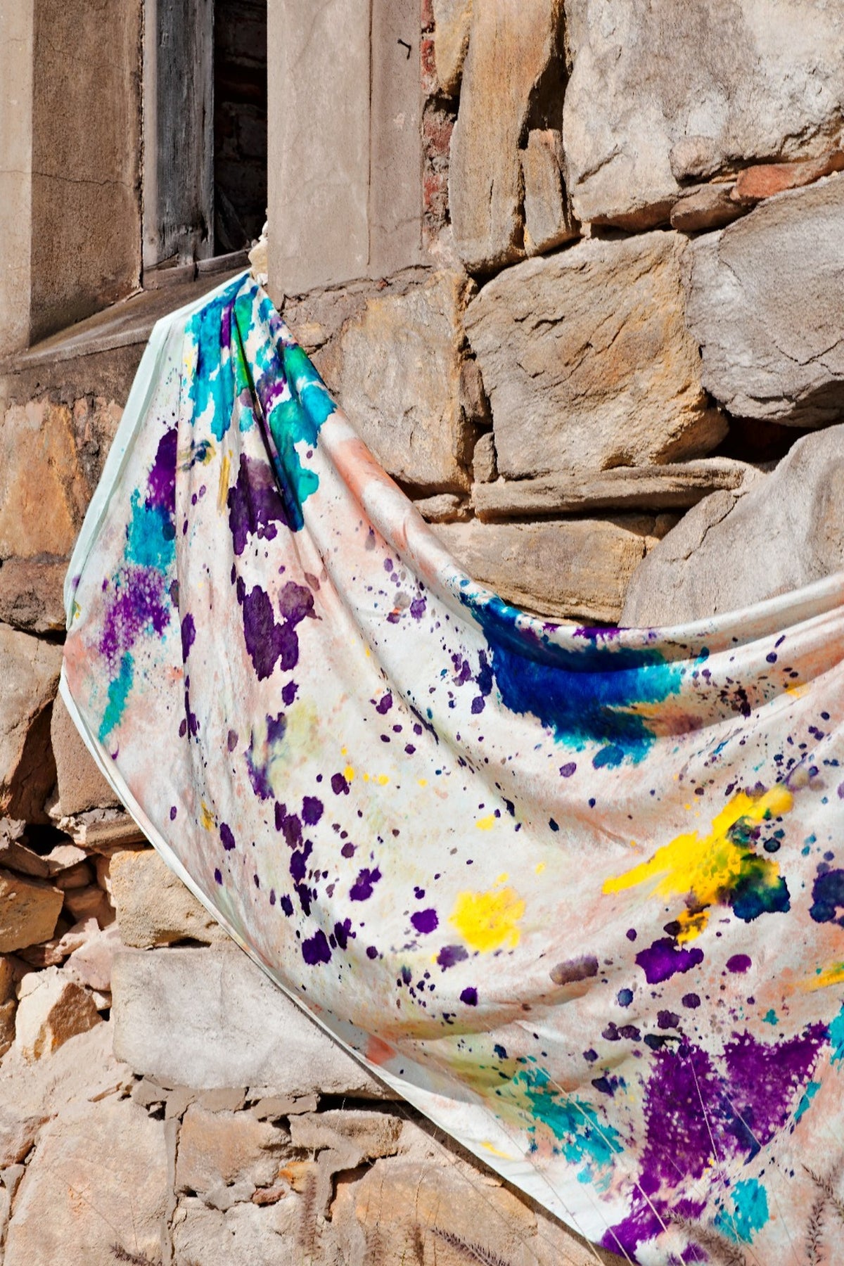 A more close up view of a hand-painted silk wrap draped on a brick wall, showcasing an abstract pattern in purple, blue, green, yellow, coral and red.