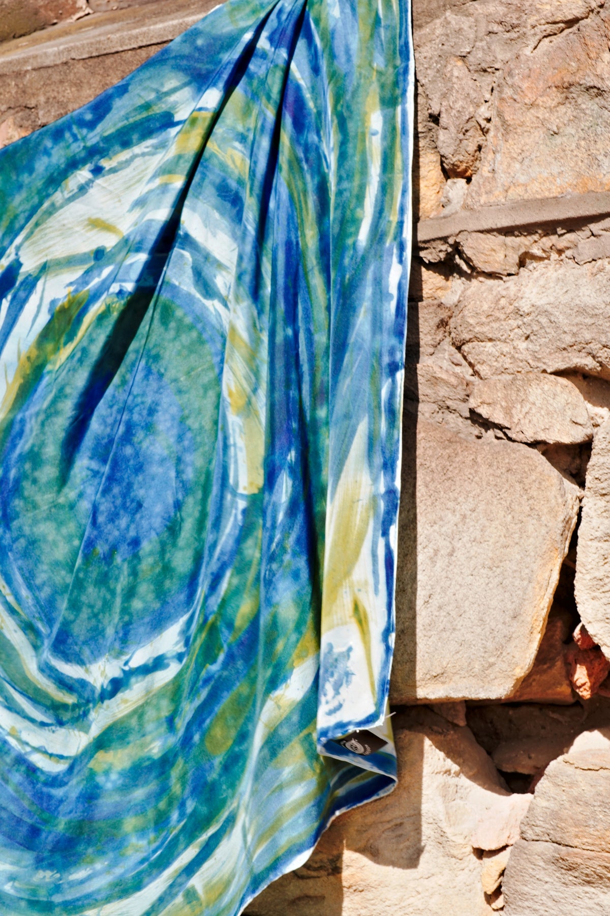 Hand-painted Mulberry silk scarf in blue, green and white with flowing abstract brushstrokes, draped against a rustic stone wall.