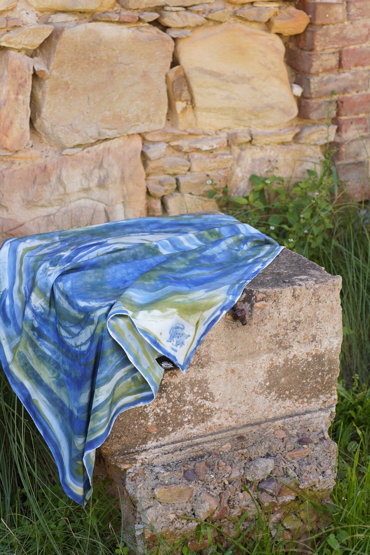 Hand-painted Mulberry silk scarf in blue, green and white with flowing abstract brushstrokes, draped over a brick stone table.