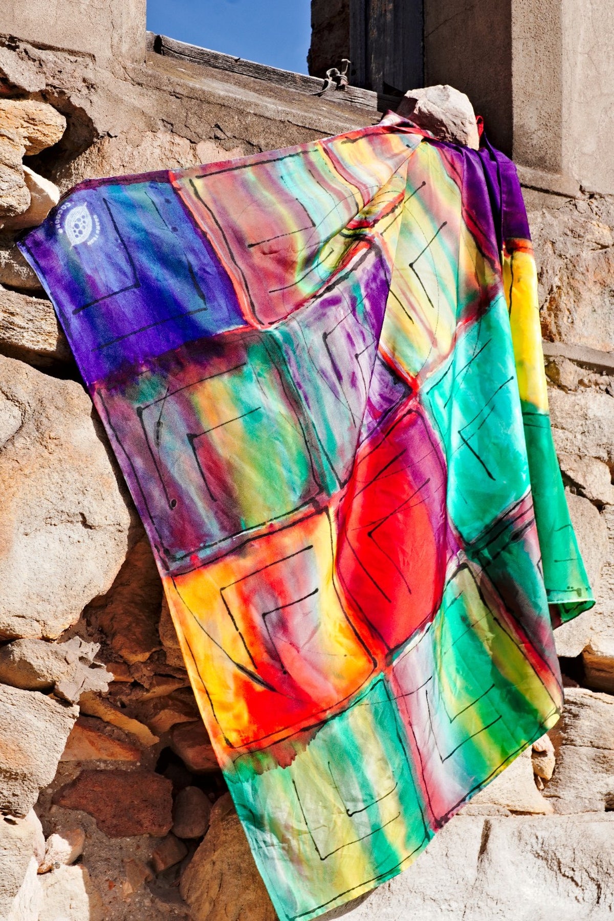 Hand-painted silk scarf draped against a rustic brick wall, showcasing an abstract colorful in purple, blue, green, yellow, red and black lines